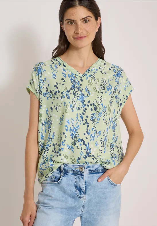 Femme portant un haut vert clair à fleurs bleues et un jean, souriante, mains dans les poches.