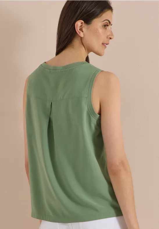 Une femme porte un top vert sans manches, vue de côté sur un fond beige.