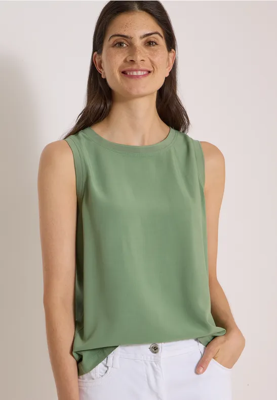 Une femme souriante porte un haut vert sans manches et un jean, posant devant un fond clair.