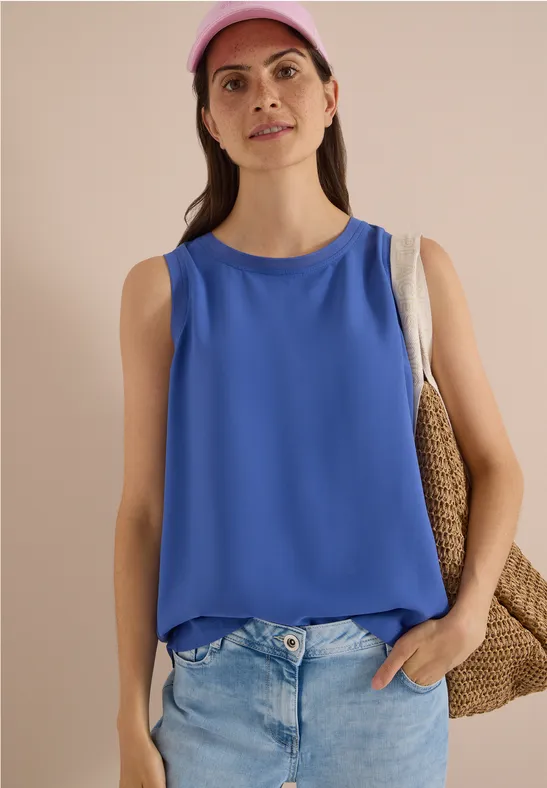 Femme en casquette rose, débardeur bleu, jean clair et sac en paille sur fond neutre.