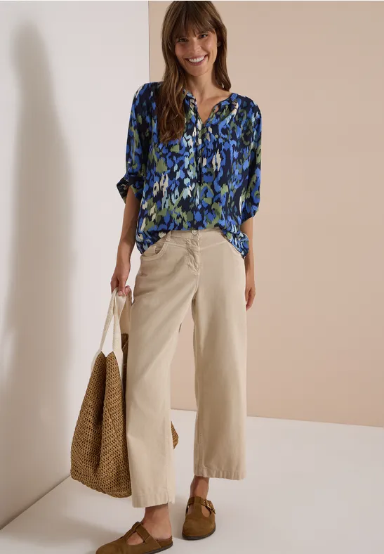 Femme souriante en chemise bleue à motifs, pantalon large beige 7/8, mocassins marron et sac en paille.