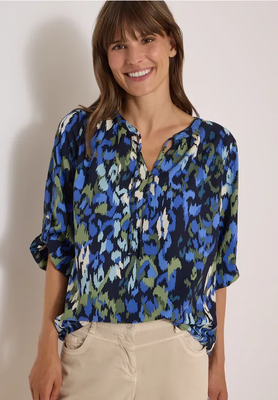 Femme souriante en chemisier bleu à motifs et pantalon beige devant un mur clair.