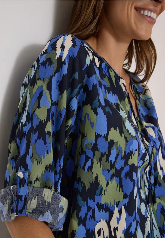 Femme de profil portant un chemisier à motifs abstraits bleu-vert avec manches retroussées.