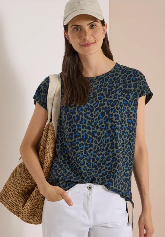 Femme en t-shirt léopard bleu-vert, pantalon blanc, sac en paille et casquette, sourire décontracté
