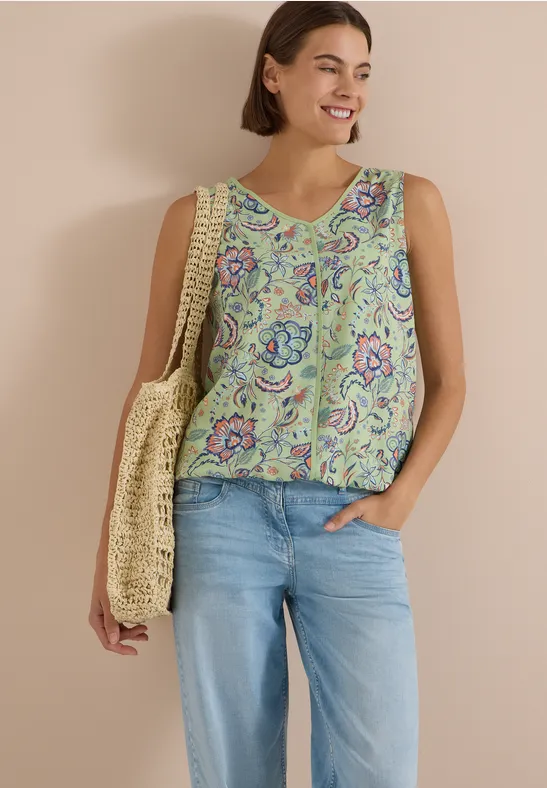 Lächelnde Frau mit ärmellosem Top im Paisley-Muster, Jeans und sommerlicher Strohtasche.