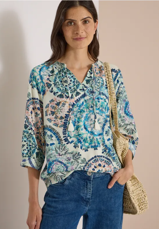 Frau in gemusterter Bluse mit Blau- und Grüntönen, Jeans und Strohtasche