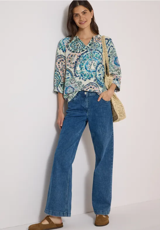 Frau in gemusterter Bluse, breiten Jeans und Stroh-Tasche vor hellem Hintergrund.