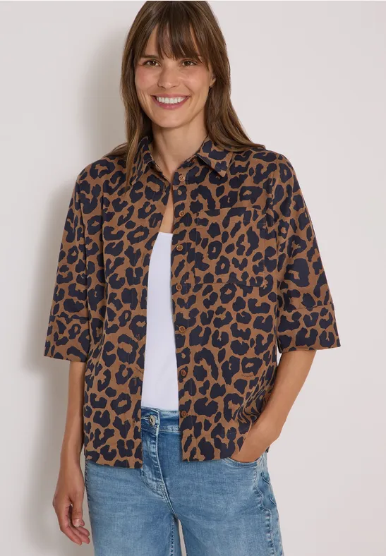 Femme souriante portant une chemise manches courtes léopard marron sur un top blanc et un jean.