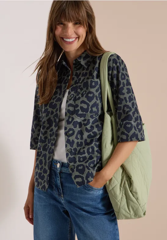 Une femme souriante porte une veste imprimée, un T-shirt blanc, un jean bleu et un sac vert.
