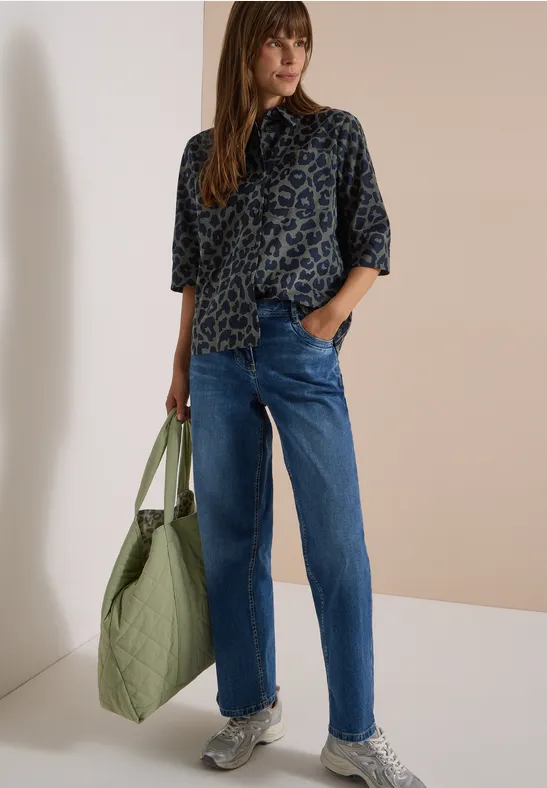 Femme en chemise léopard, jean bleu, baskets, tient un grand sac vert
