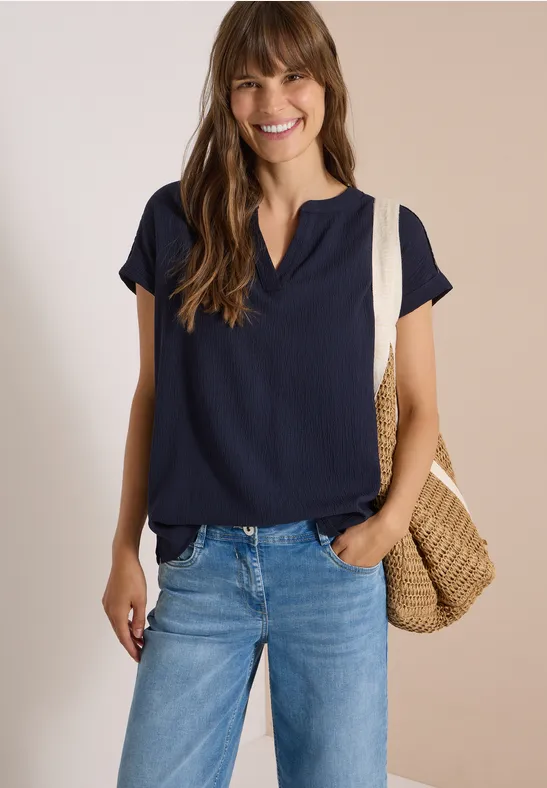 Lächelnde Frau trägt dunkelblaue Bluse, Jeans und eine geflochtene Strohtasche.