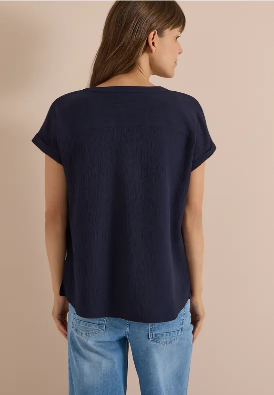 Frau steht Rücken zur Kamera, trägt dunkelblaues T-Shirt mit kurzen Ärmeln und helle Jeans.