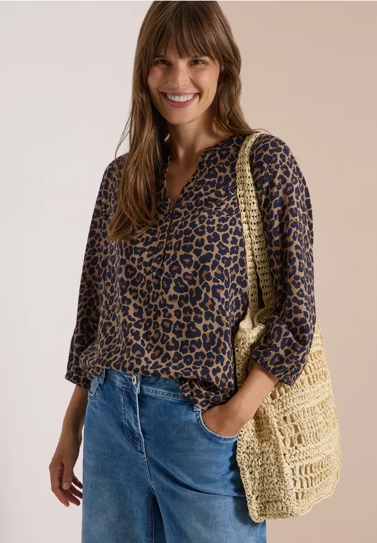 Femme souriante portant chemise léopard, jean bleu et sac tressé.
