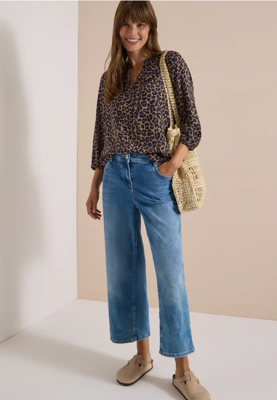 Femme portant un chemisier léopard, jean large court, espadrilles et sac tressé sur fond clair.