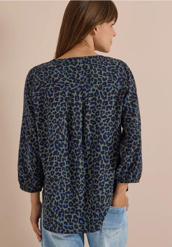 Frau von hinten in locker sitzender Bluse mit blau-grünem Leopardenmuster und hellen Jeans.
