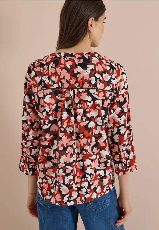 Femme de dos portant une blouse fleurie rouge, blanc et noir à manches trois-quarts, associée à un jean bleu.