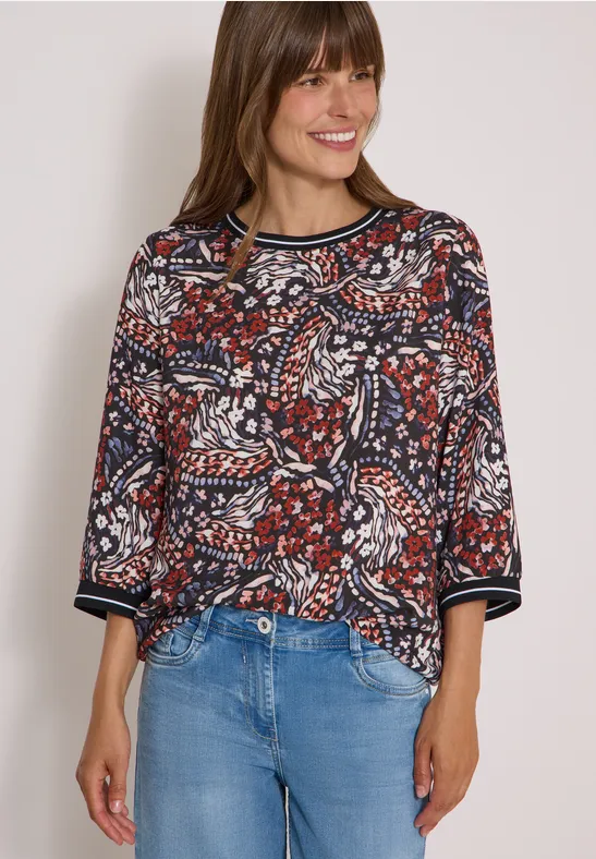 Femme souriante en blouse à motifs colorés et jean devant un fond clair.