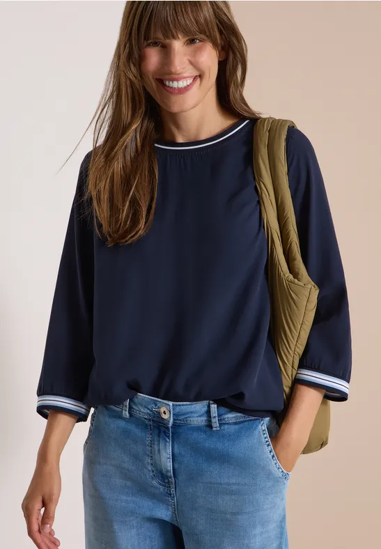 Femme souriante en chemisier bleu marine à rayures blanches au col et aux manches, jean et sac matelassé vert.