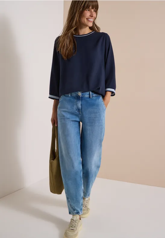 Femme en pull bleu et jean, sac à l'épaule et baskets sur fond beige.