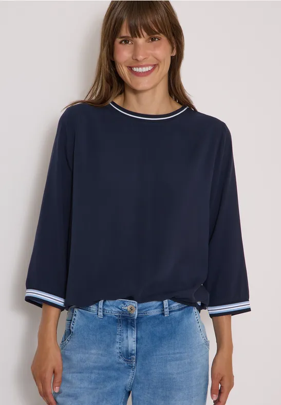 Femme souriante en chemise bleu marine et jean devant un fond clair.