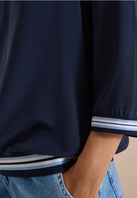 Gros plan d’une personne en veste de sport bleu foncé à poignets rayés, jeans bleus, main dans la poche.