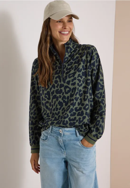 Une femme souriante porte une veste polaire vert foncé à motif noir, un jean et une casquette grise.