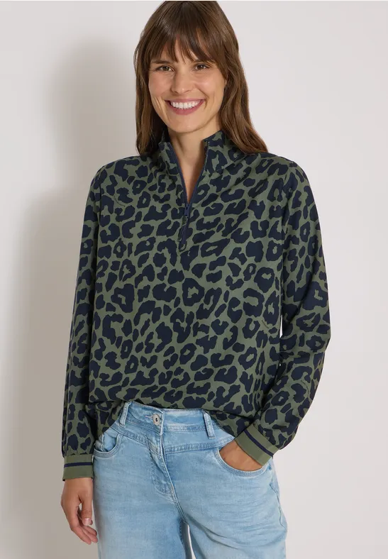 Une femme souriante porte un pull vert à motif léopard et un jean bleu devant un fond clair.
