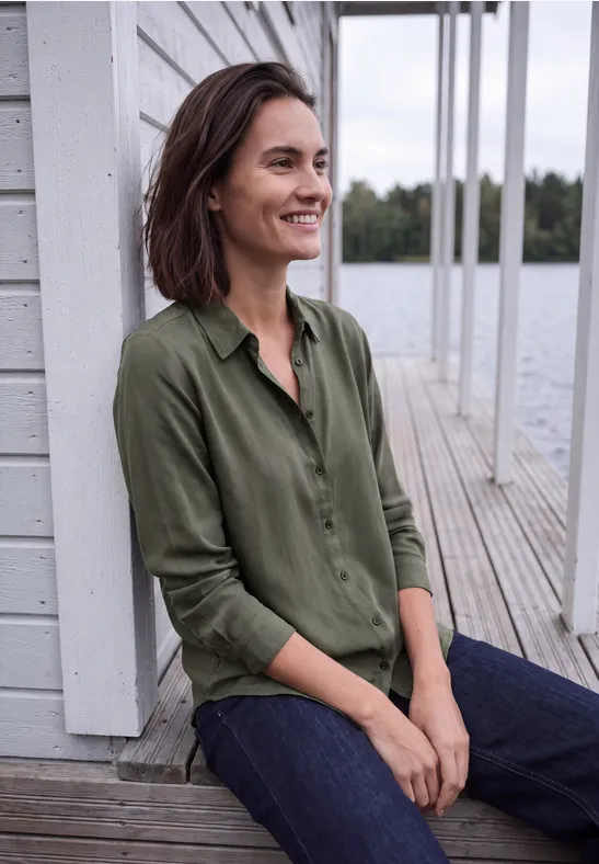 Une femme souriante en chemise verte est assise paisiblement sur une jetée en bois au bord de l'eau.