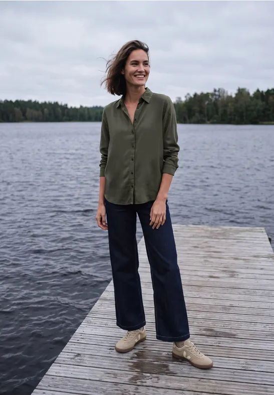 Une femme sur un ponton en bois au bord d’un lac, portant une chemise verte, un jean et des chaussures claires.