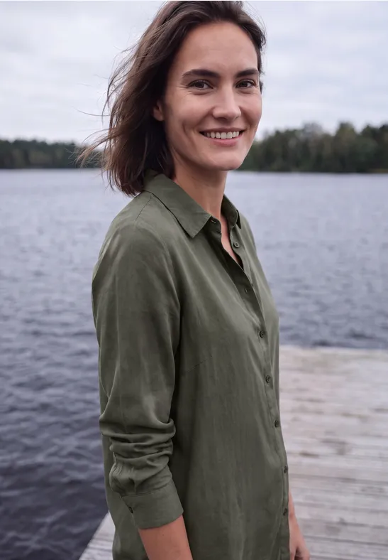 Une femme souriante se tient sur une jetée au bord d’un lac, vêtue d’une chemise verte et regarde l’objectif.
