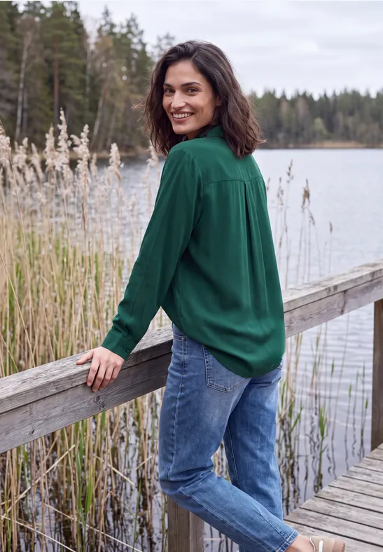 Frau im grünen Hemd lehnt lächelnd am Holzsteg an einem See, umgeben von Schilf und Wald im Hintergrund.