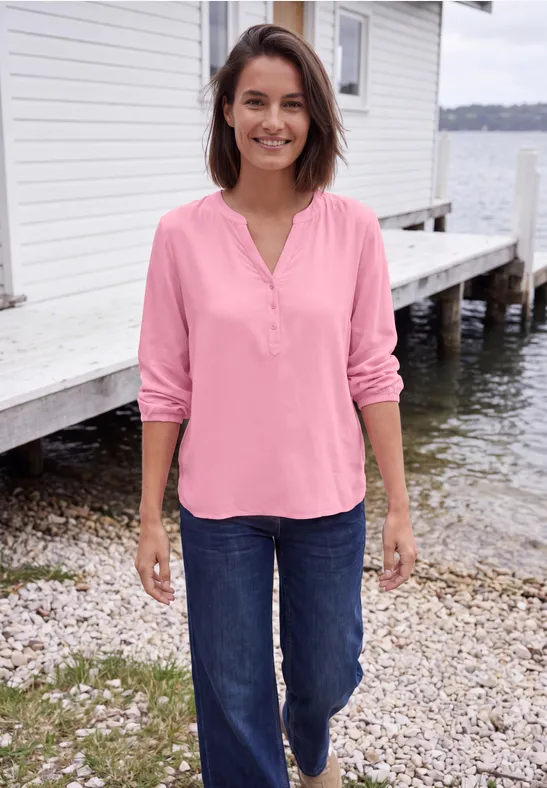 Femme souriante portant un chemisier rose et un jean bleu, debout près d'une cabane au bord de l'eau.