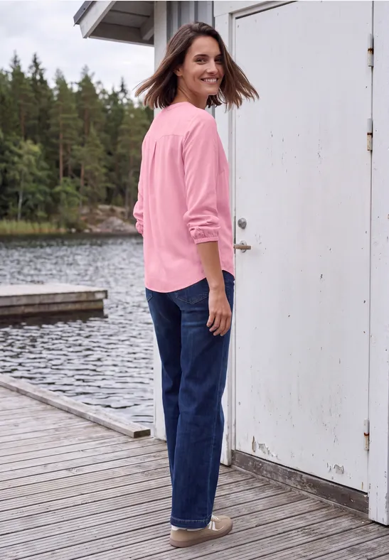 Une femme en pull rose et jean se tient sur un ponton au bord du lac, regardant en arrière et souriant.