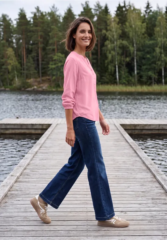 Une femme en pull rose et jean marche avec le sourire sur une jetée en bois au-dessus d'un lac tranquille.