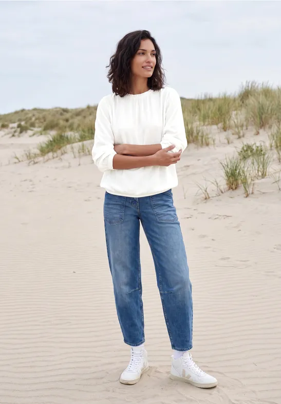 Frau in weißem Pullover, blauen Jeans und weißen Sneakern steht lächelnd mit verschränkten Armen am Sandstrand.