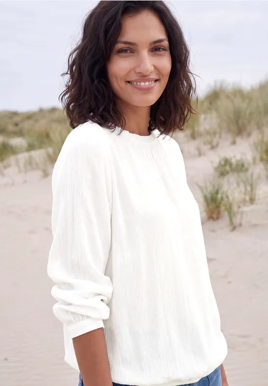 Lächelnde Frau im weißen Pullover steht am sandigen Strand mit Dünen im Hintergrund.
