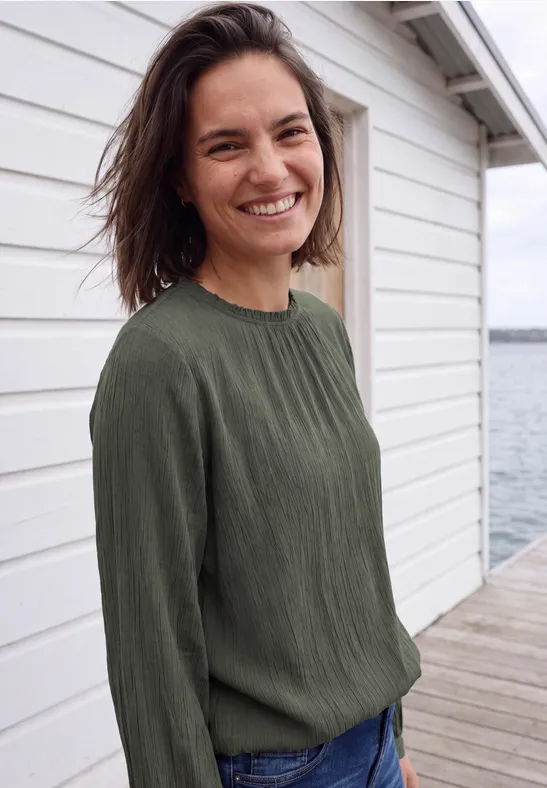 Une femme souriante aux cheveux bruns, vêtue d'un haut vert, debout sur une jetée devant une cabane blanche.