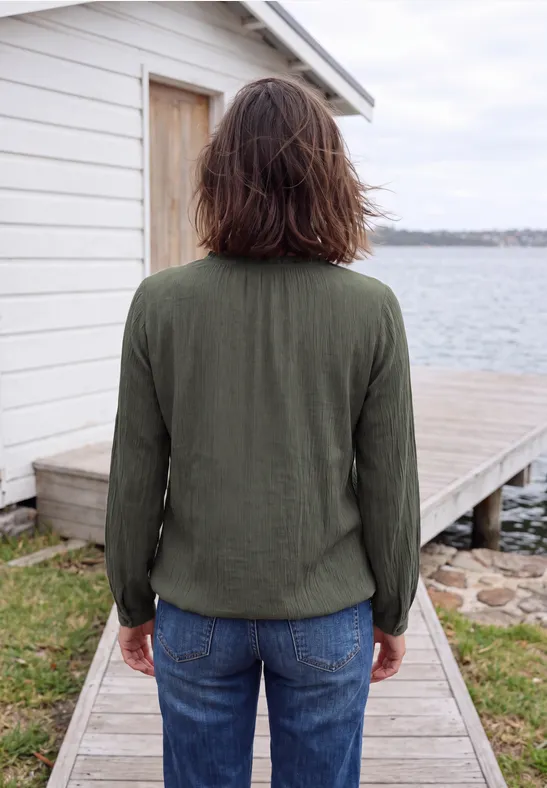 Personne en veste verte et jean, debout sur une passerelle en bois devant une cabane blanche au bord de l'eau.