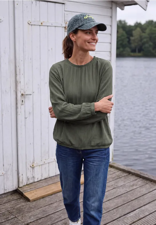 Femme en casquette et haut vert, bras croisés, sur un ponton en bois devant un lac.