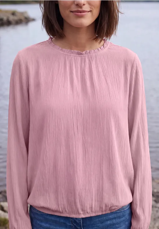 Frau in rosa Bluse, stehend vor einem See, Oberkörper von Kinn bis Taille sichtbar