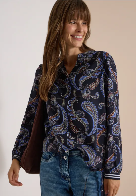 Frau mit langen braunen Haaren lächelt, trägt dunkle Paisley-Bluse und Jeans.