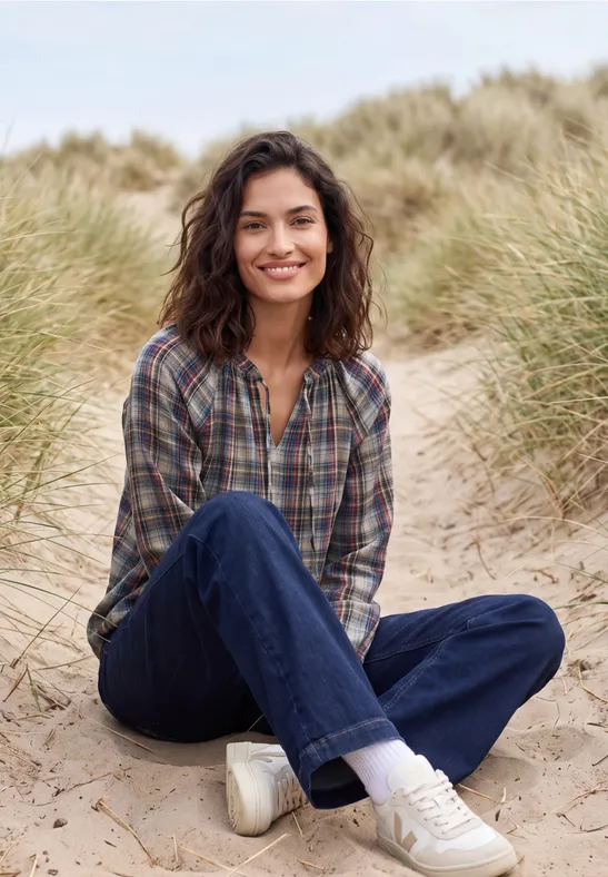 Frau sitzt lächelnd im Sand vor Dünen, trägt kariertes Hemd, Jeans und weiße Sneaker.