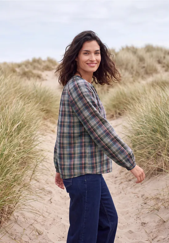 Eine lächelnde Frau in kariertem Hemd und Jeans wandert über sandige Dünen mit Gras.