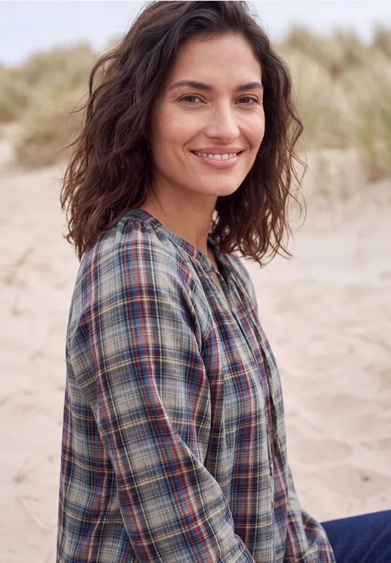 Eine lächelnde Frau mit dunklen, lockigen Haaren in einem karierten Hemd steht am Strand mit Dünen im Hintergrund.