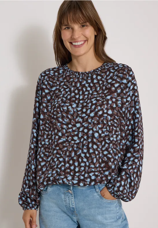 Jeune femme souriante en chemisier à motifs et jean, devant un fond clair.