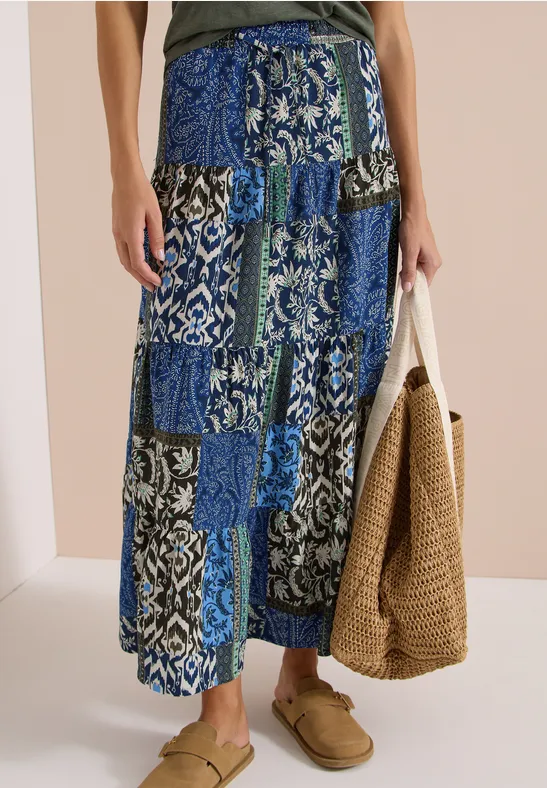 Maxirock mit blau-weißem Patchwork-Muster, locker sitzend, kombiniert mit Strohtasche und Hausschuhen.