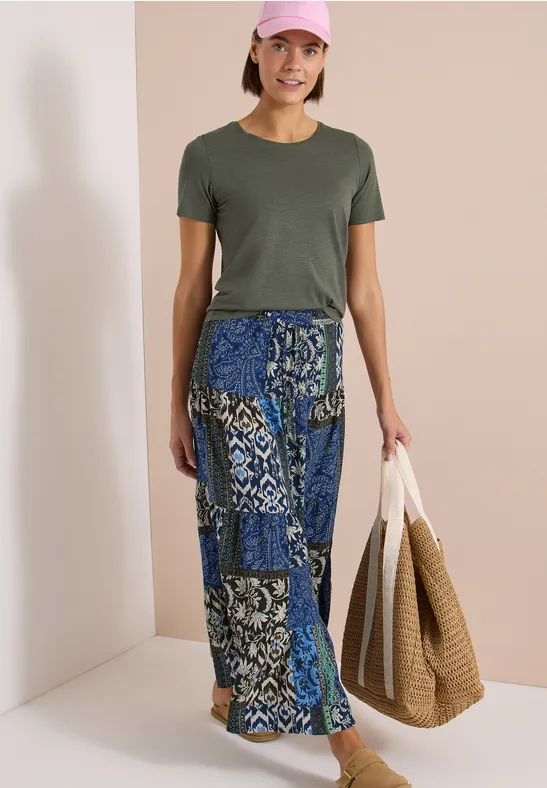 Person in olivgrünem T-Shirt und langem blau gemustertem Rock hält zwei große Taschen.