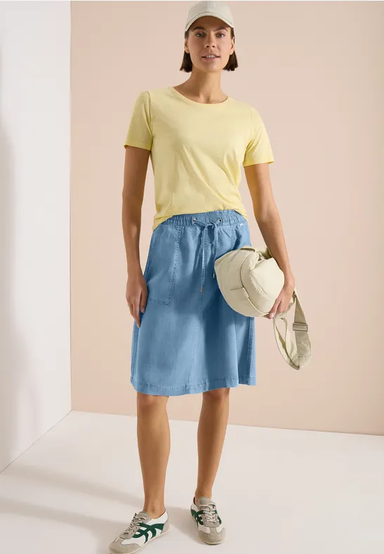 Frau in gelbem T-Shirt, leichtem Jeansrock, Sneakern, Cap und Tasche vor hellem Hintergrund.