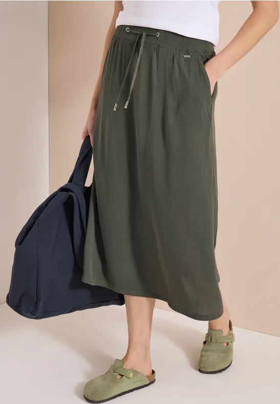 Frau trägt olivgrünen Midirock mit Tunnelzug, weißem Top, grünen Sandalen und blauer Tasche.