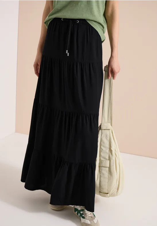 Une personne porte une jupe longue noire à volants, un haut vert, un sac beige et des baskets vertes.